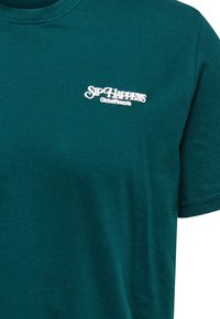 Camiseta de algodón color azul verdoso con mangas cortas y cuello redondo. En la parte frontal lleva bordado el logo "Sip Happens GlobalResorts" en blanco.