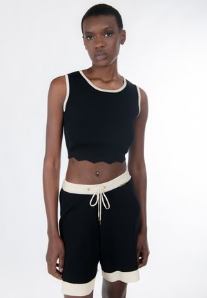 Personne mince portant un haut court noir sans manches et un short assorti avec un bord beige et un cordon de serrage, debout devant un fond blanc.