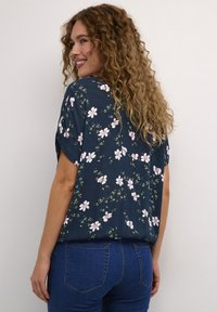 Donna con capelli ricci sorride, indossando una blusa corta a maniche con fiori di colore blu scuro e jeans blu, in piedi contro uno sfondo chiaro e semplice.