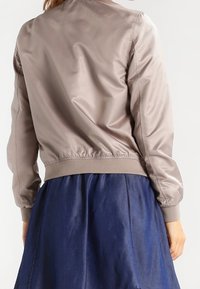 Lichte taupe bomberjack met een soepele, glanzende textuur, elastische tailleband en ribbelmanchetten, gecombineerd met een marineblauwe rok.