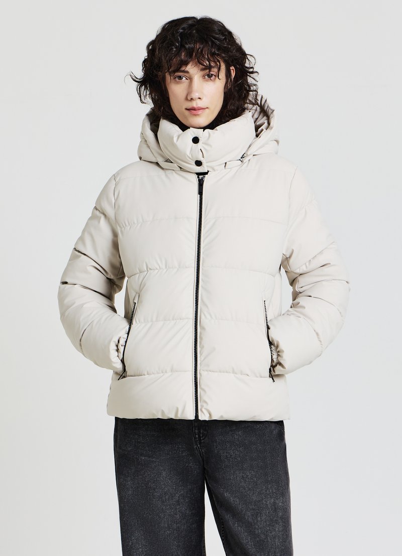 Giacca imbottita beige con cappuccio, dotata di cerniera frontale, due tasche laterali e polsini elastici. Tessuto liscio con design trapuntato.