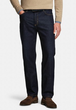 Homme portant un jean droit bleu foncé avec une ceinture marron et des mocassins en daim marron, pull beige sur une chemise bleue, mains détendues.