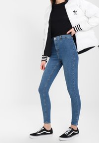 Jeans ajustados de mezclilla azul con una cintura alta, combinados con una camiseta negra, una chaqueta blanca acolchada y zapatillas negras con suelas blancas.