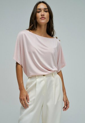 Femme portant un chemisier rose clair à manches courtes avec des boutons sur une épaule et un pantalon blanc taille haute à jambes larges.