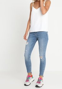 Bílý bezrukávový top s výstřihem do V, spárovaný s pastelově modrými skinny džíny s oprýskanými detaily a barevnými teniskami.