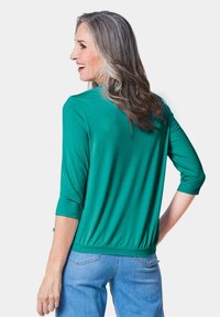 Teal 3/4 mouw top met een gathered onderrand, een gladde textuur en een subtiele glans. Gecombineerd met lichtblauwe jeans.
