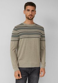Man in een beige trui met zwarte, witte en grijze geometrische patronen op de borst en schouders, gecombineerd met donkere jeans.