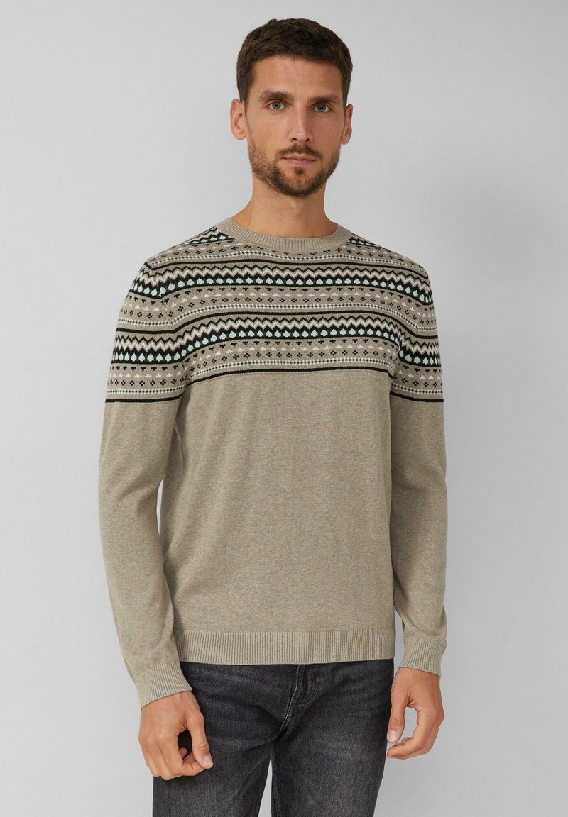 Man in een beige trui met zwarte, witte en grijze geometrische patronen op de borst en schouders, gecombineerd met donkere jeans.