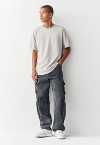 Harmaa puuvillainen t-paita ja tummanharmaat taskupussilliset cargo-housut, joissa on teksturoitu kangas, yhdistettynä valkoisiin lenkkareihin. Seisoma-asento valkoista taustaa vasten.