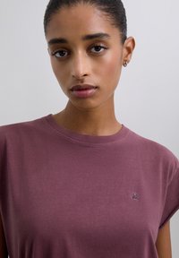 Mujer joven con el cabello liso recogido, usando un pequeño aro en la oreja y una camiseta sin mangas de color malva con un pequeño logo de calavera en el pecho.