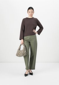 Kvinne iført brun langermet topp, olivengrønne bukser, svarte spisse sko, som holder en beige håndveske med slangeskinnmønster, står på hvit bakgrunn.
