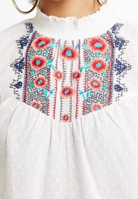 Vit blus med en rynkad halsringning, prydd med intrikata blommiga broderier i rött, blått och grönt, i kontrast till en strukturerad tyg.