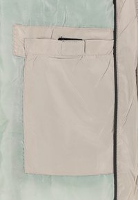 Veste isolante vert clair avec une texture lisse et brillante ; elle présente une poche beige avec une fermeture éclair noire et des détails de coutures contrastantes.