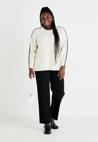 Chunky Creme-Pullover mit strukturiertem Strick, schwarzen Seitenelementen; schwarze weit geschnittene Hose; schwarze Stiefeletten mit Absatz; hält eine kleine weiße Tasche.