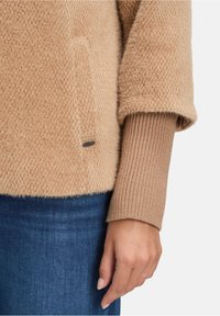 Ręka spoczywająca obok jasnobrązowej, teksturowanej wełnianej kurtki z prążkowanym mankietem, noszonej na niebieskich dżinsach na białym tle.