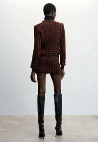Giacca su misura in rosso e nero a motivo houndstooth con orli frangiati e maniche lunghe, abbinata a una mini gonna coordinata e stivali alti neri con tacco a spillo.