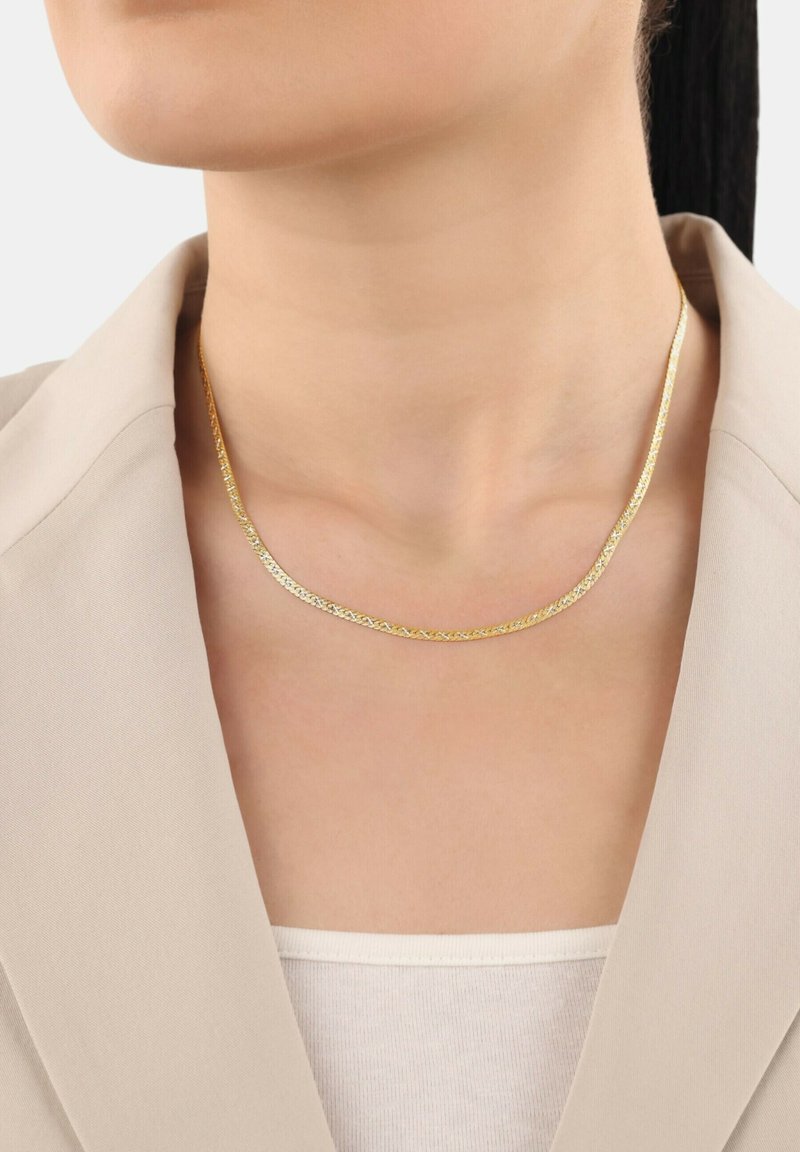 Guldhalsband med en ömtålig, vriden design. Bärs på en bar nacke, tillsammans med en ljusfärgad blazer och topp.