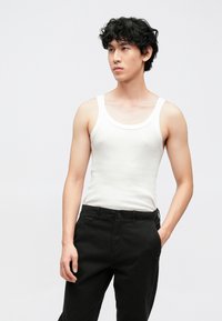 White cotton tank top with a round neckline and wide straps, paired with black cotton pants. Model stands with one hand in pocket.