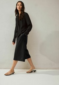 Must mustrindiga must blouse, millel on sidumiseelne kaelus, koos musta põlvepikkuse seeliku ja mustade rihmadega sandaalidega, millel on tekstuurne viimistlus.