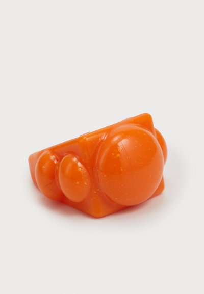 Pomarańczowy plastikowy pierścień o geometrycznym wzorze, z trzema wystającymi okrągłymi kształtami o różnych rozmiarach, gładką teksturą i błyszczącym wykończeniem.