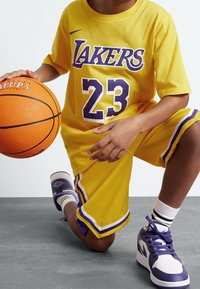 Keltainen koripallopaita, jossa on "Lakers" ja "23" purppuralla, yhdistettynä samansävyisiin shortseihin ja valkoisiin tennoihin purppuravihjeillä. Koripallo kädessä.