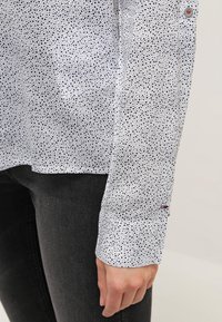 Biała koszula w czarne kropki, z długimi rękawami i guzikiem na mankiecie z czerwonymi i niebieskimi akcentami, pokazana obok ciemnoszarych spodni.