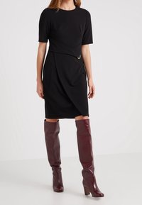 Svart klänning med en figurnära bodice, korta ärmar och draperad front, accentuerad med en guldring. Matchad med höga, glänsande burgunderröda knästövlar.