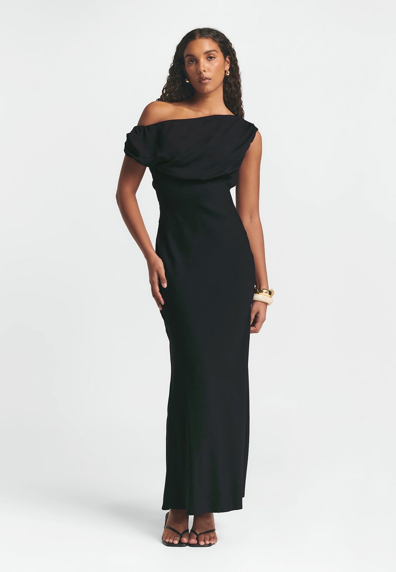 Juoda su peties nuleista maksi suknelė su drapiruota medžiaga, prisitaikanti siluetas ir lygus paviršius, derinama su paprastais juodais sandalais.