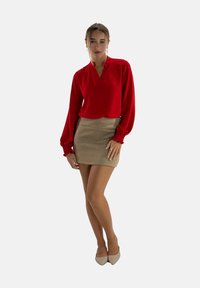 Blusa rossa a maniche lunghe con scollo a V e polsini arricciati abbinata a una mini gonna color marrone chiaro. Modella in tacchi beige, in piedi contro uno sfondo bianco.