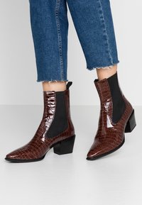 Botas de tobillo de imitación de cocodrilo marrón con paneles laterales elásticos, puntera afilada y tacón cuadrado, combinadas con unos jeans de denim azul cortos.