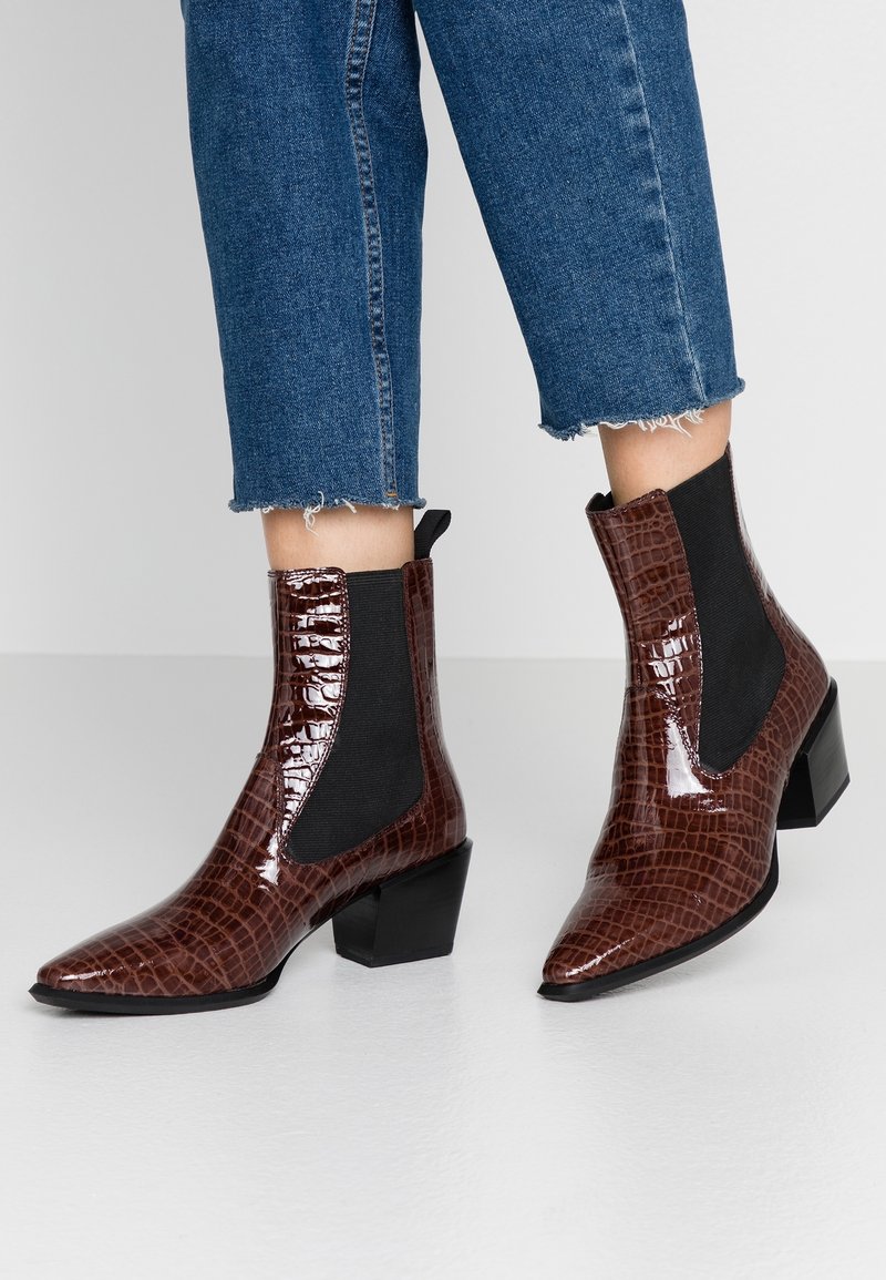 Botas de tobillo de imitación de cocodrilo marrón con paneles laterales elásticos, puntera afilada y tacón cuadrado, combinadas con unos jeans de denim azul cortos.