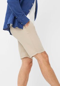 Pantaloni beige realizzati in tessuto leggero, con design a gamba dritta, tasche laterali e una superficie liscia. Abbinati a una camicia blu.