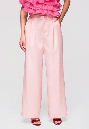 Vrouw met hoge taille, wijde roze broek, roze top met ruches en rode open-teen hakken, staand met één hand in haar zak.