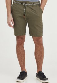 Shorts vert olive en tissu texturé, avec une ceinture tressée, des passants de ceinture et des poches latérales. Portés avec des baskets noires.