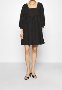 Svart kort klänning med fyrkantig halsringning, puffärmar och lätt texturerad tyg; bärs med svarta öppna klackar.