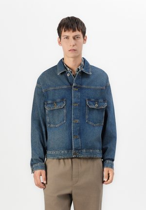 Giovane uomo che indossa una giacca di jeans blu sopra una camicia a quadri con pantaloni beigi, in piedi contro uno sfondo bianco semplice.