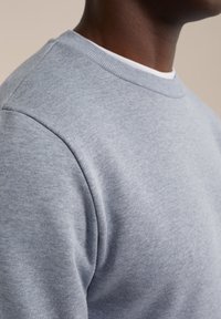 Närbild av en person som bär en ljusgrå sweatshirt med rund hals över en vit t-shirt, på en neutral bakgrund.