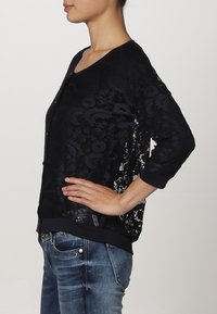 Cardigan in pizzo nero con un motivo floreale, maniche 3/4 e orlo a contrasto. Indossato sopra una maglietta semplice e abbinato a jeans blu.