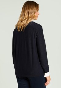 Granatowy dzianinowy sweter z pionowym prążkowaniem; luźna sylwetka, długie rękawy i biała koszula z kołnierzykiem widoczna przy mankietach.