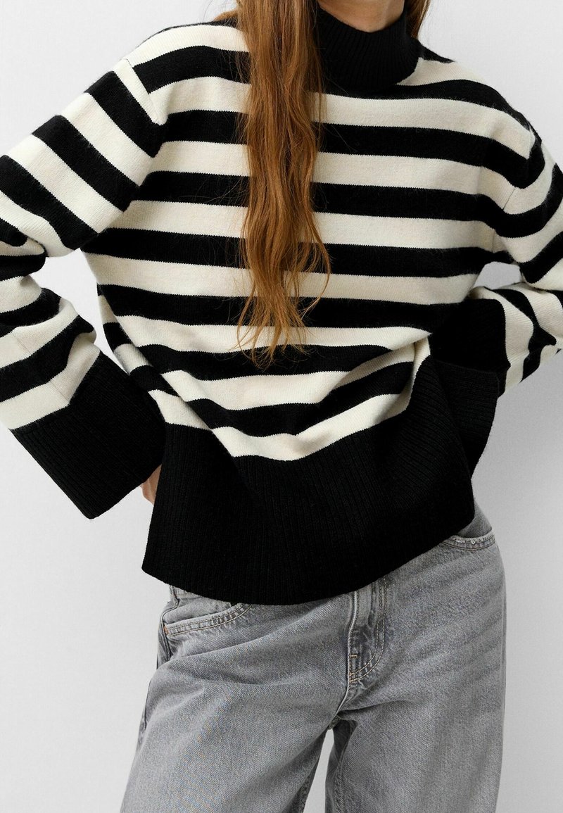 Personne portant un pull rayé horizontal noir et blanc à manches longues et un jean gris clair, debout les mains sur les hanches.