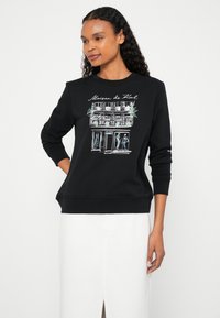 Čierna mikina s potlačou obchodíka s nápisom „Maison de Korl“, jemnými detailmi a rebrovanými manžetami a lemom.