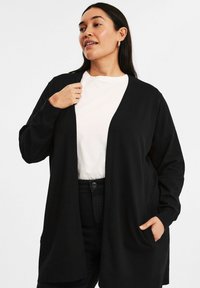 Svart cardigan med avslappnad passform, öppen front, långa ärmar och sidofickor, bärs över en vit skjorta och svarta jeans. Mjuk textur.