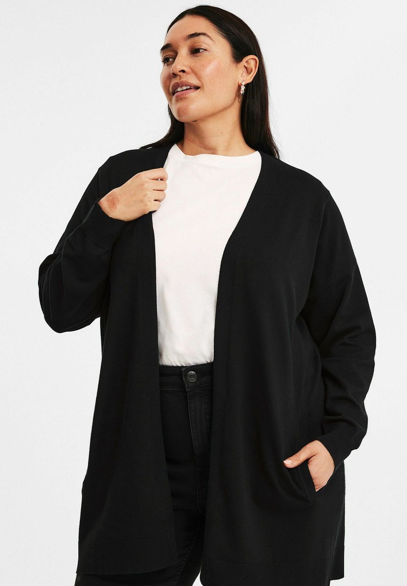 Svart cardigan med avslappnad passform, öppen front, långa ärmar och sidofickor, bärs över en vit skjorta och svarta jeans. Mjuk textur.