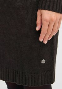Gebreide donkerbruine sweaterjurk met geribbelde onderrand. Metaal logo detail aan de zijkant. De stof lijkt zacht en textuur te hebben.