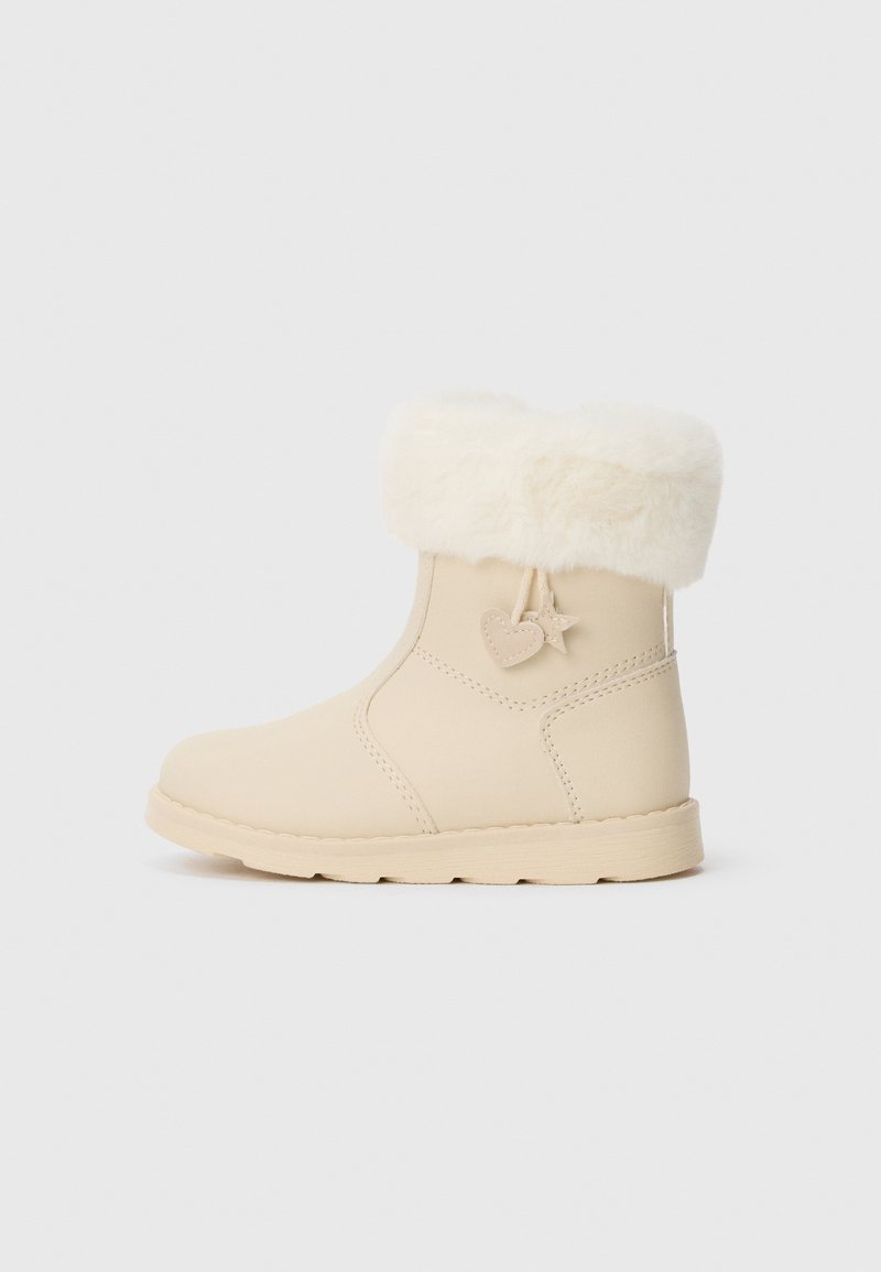 Botte d’hiver couleur crème avec bord en fausse fourrure, couture latérale, et breloques en forme de cœur et d’étoile suspendues à la cheville extérieure.