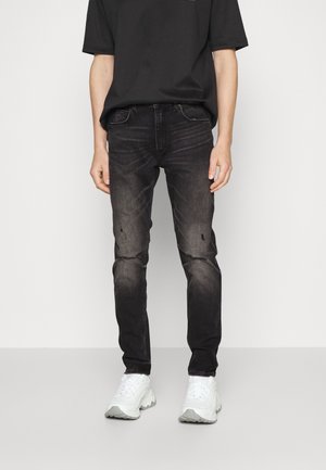 Henkilö, joka käyttää mustia kuluneita kapeita farkkuja, joissa on polvivaurioita, ja valkoisia paksupohjaisia lenkkareita, seisoo yksiväristä taustaa vasten.