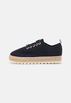 Czarne dzianinowe sneakersy z midsole o teksturze liny, białą zewnętrzną podeszwą z rowkami, czarnymi sznurowadłami i złotymi oczkami na białym tle.
