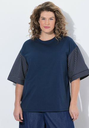 Frau mit lockigem Haar, die ein marineblaues Hemd mit breiten Nadelstreifenärmeln und blaue Hosen trägt, steht vor einem hellen Hintergrund.