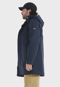 Parka impermeabile blu navy con cappuccio, tasche frontali e toppa con logo sulla manica sinistra. Presenta un orlo dritto e polsini con bottoni.