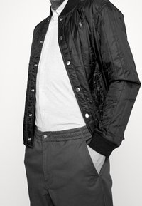 Svart kviltad bomberjacka med silverfärgade tryckknappar, ribbade ärmslut och en liten logotyp på bröstet, matchad med en ljusgrå T-shirt och grå byxor.